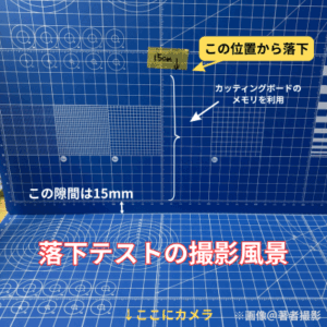 ガンプラパーツの落下テストの撮影風景の画像。青いカッティングマットを２枚使っている。一枚を立てて、１５c mの位置にマスキングテープで印をつけ、カッティングボードのメモリで跳ね返り高さがわかりやすくしている。