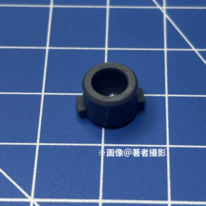 ガンプラのパーツ。跳ね返り実験に用いたピース。素材はP E、大きい方。筒状の底部分の0度と180度の位置に四角い突起がある。濃いグレー